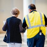 Profissional de segurança auxilia uma mulher idosa no corredor de um prédio moderno.