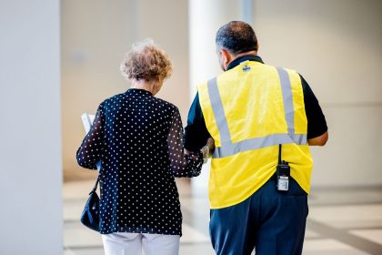 Profissional de segurança auxilia uma mulher idosa no corredor de um prédio moderno.